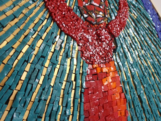 The image is a square mosaic piece, set against a neutral, off-white background. The artwork itself is diamond-shaped in orientation, with the corners of the square pointing upwards, downwards, and sideways. The artwork features a central figure with arms raised, holding a golden orb, all encompassed within concentric circular and square borders.
