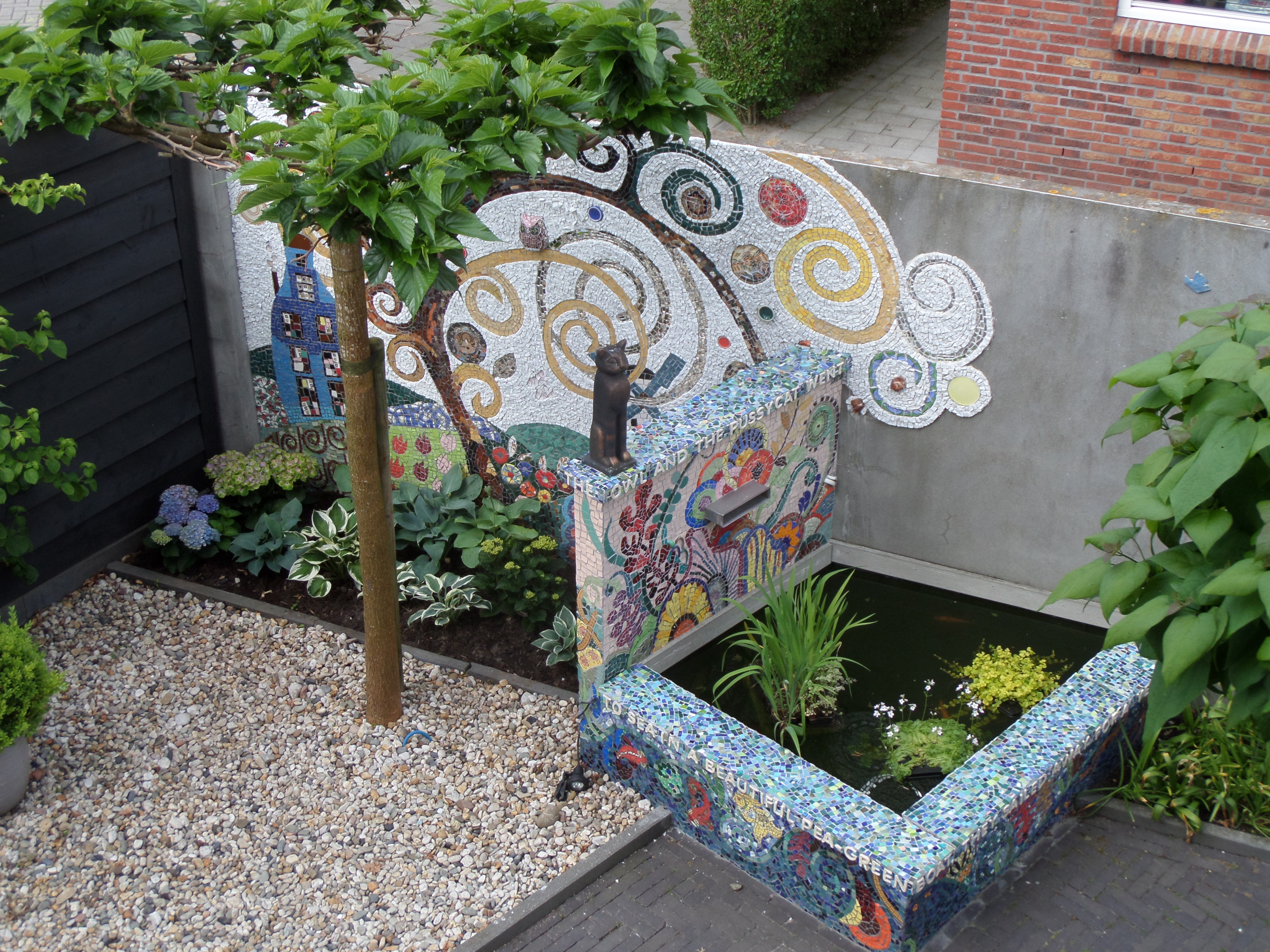 The main focus is a small, creatively designed garden space. In a niche or corner adjacent to a home. A prominent feature is a raised planter or water feature decorated with mosaic tiles. Next to this, we can see tree with a thin trunk. The other part is a wall decorated with another mosaic.