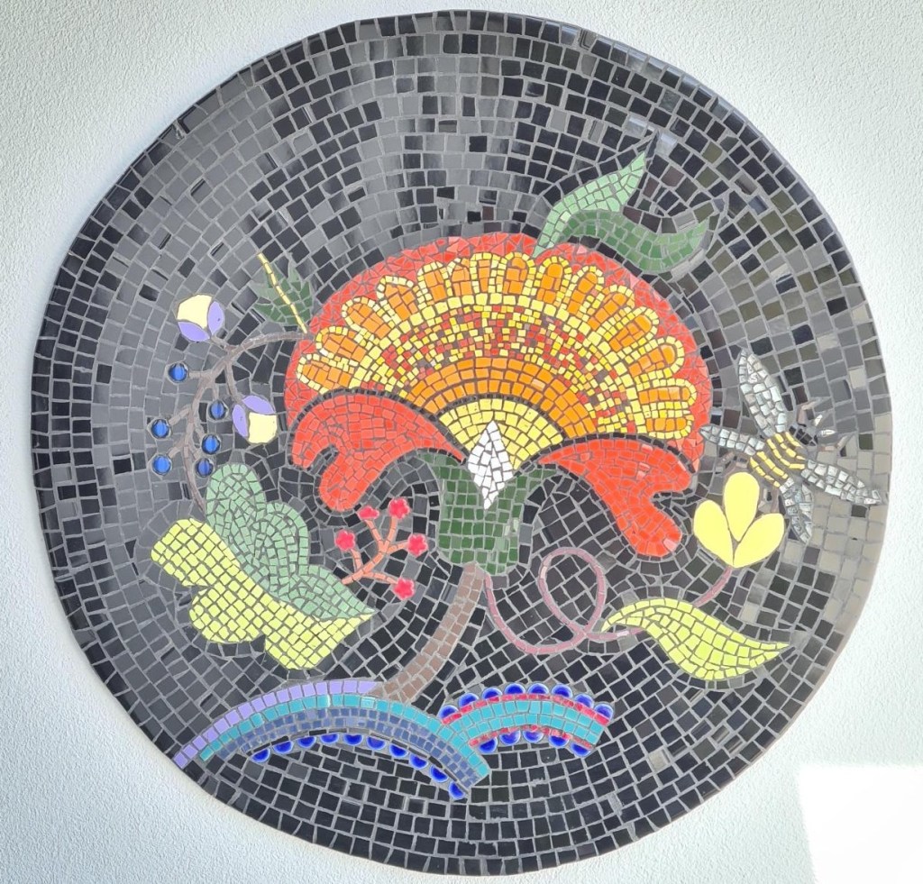 A vibrant mosaic artwork is centrally positioned within the frame, displaying an ornate floral design. The artwork is circular, with a border composed of small, dark gray, rectangular tiles. The central design features a large, stylized flower in shades of orange, red, and yellow, with a white center and detailed, petal-like structures. Surrounding the main flower are various botanical elements, including green leaves, blue berries, small yellow flowers, and a detailed insect that resembles a dragonfly or a bee. These elements are rendered in shades of green, yellow, and blue, creating a vibrant contrast against the dark background. The background has a pattern of dark grey squares. The artwork is attached to a plain, off-white wall. The lighting appears soft and even, highlighting the intricate details of the mosaic. The perspective is straightforward, showcasing the artwork directly. The overall style is artistic and decorative, with a strong emphasis on color and detail, creating a sense of craftsmanship and visual interest.