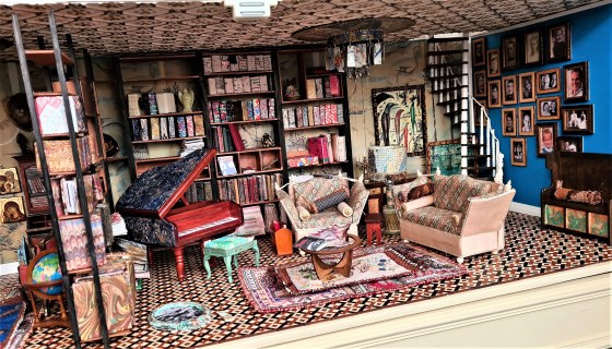 The image shows an intricately designed miniature room of a dollhouse interior. It's set up as a cozy and cluttered living space, filled with books, furniture, and various decorative items. The overall impression is one of a well-loved and lived-in room, suggesting a comfortable and creative atmosphere. Walls: The walls are lined with tall bookshelves, filled with what appear to be miniature books. One wall is painted a bold blue color, contrasting with the warmer tones of the rest of the room. There's a collection of small framed pictures on the blue wall.