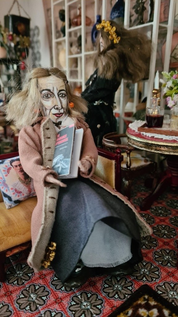 The doll in the foreground is seated on a small, yellow cushioned chair. She is wearing a long, light-pink coat with beige trim, a dark-blue or grey skirt, and seems to be holding a small book or booklet. She has long, slightly disheveled blonde hair.