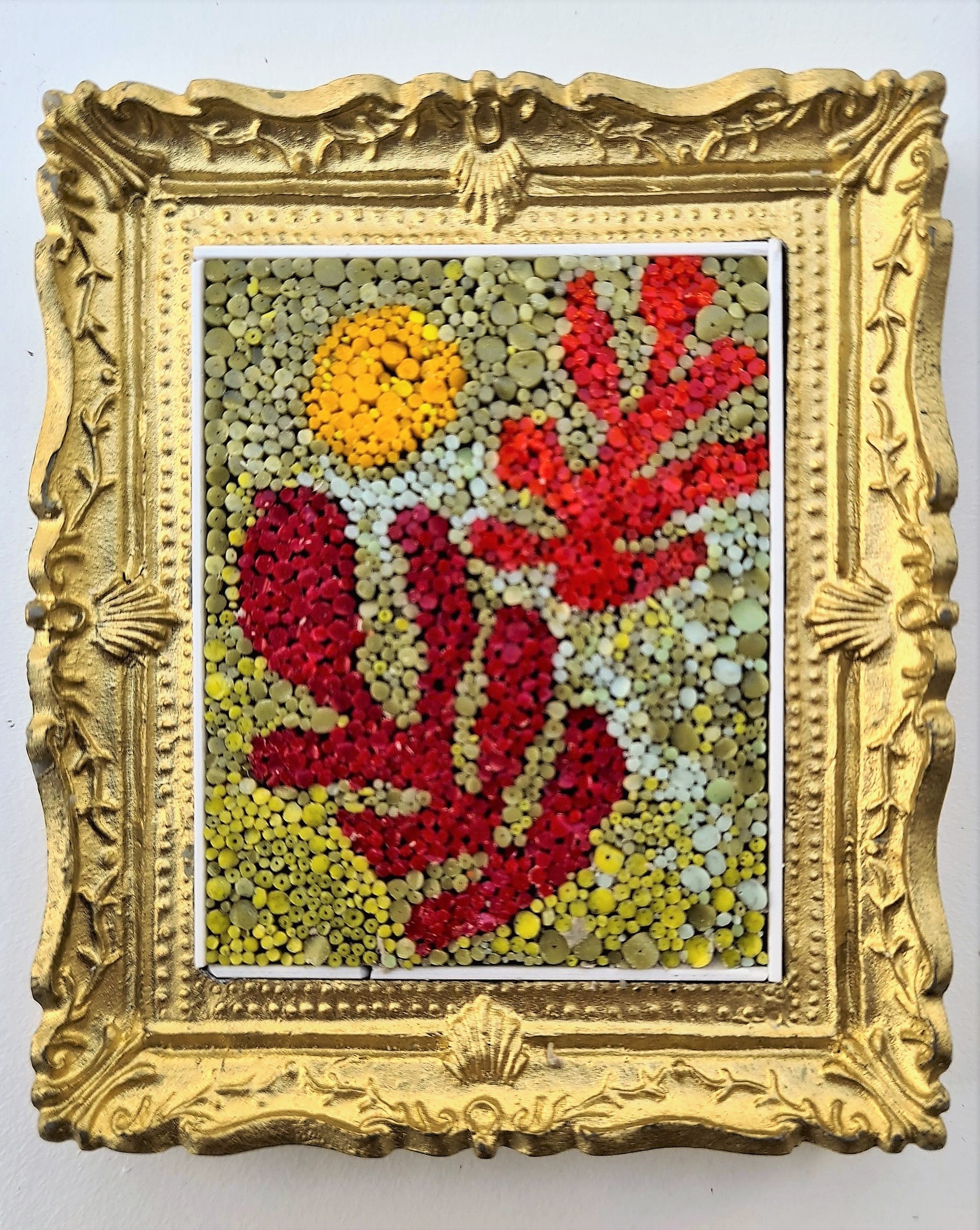 The image is of a framed artwork. The artwork itself appears to be a mosaic or collage composed of small, colorful elements. The subject matter is somewhat abstract, reminiscent of organic forms or stylized plants. There is a prominent circular yellow shape in the upper left, suggesting a sun or moon. Dominating the composition are red shapes that could be interpreted as leaves, coral, or other natural forms.