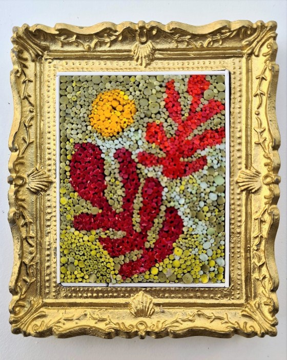 The image is of a framed artwork. The artwork itself appears to be a mosaic or collage composed of small, colorful elements. The subject matter is somewhat abstract, reminiscent of organic forms or stylized plants. There is a prominent circular yellow shape in the upper left, suggesting a sun or moon. Dominating the composition are red shapes that could be interpreted as leaves, coral, or other natural forms.